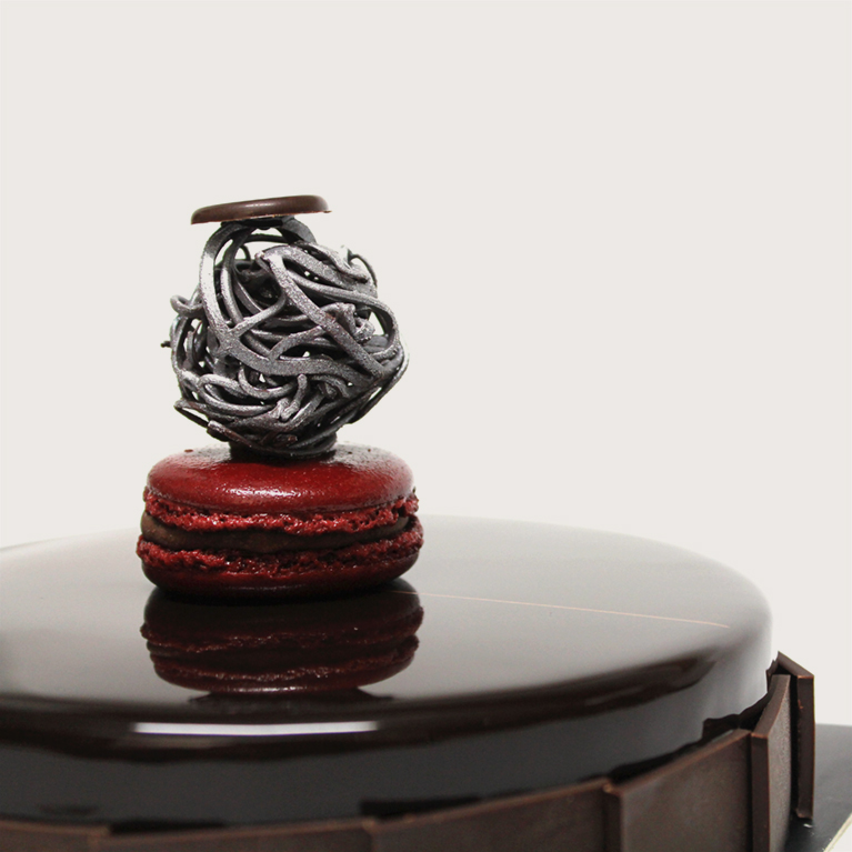 A red French macaron with a silver ornament and a piece of chocolate placed on top of a chocolate cake that has chocolate pieces on its rounds.
