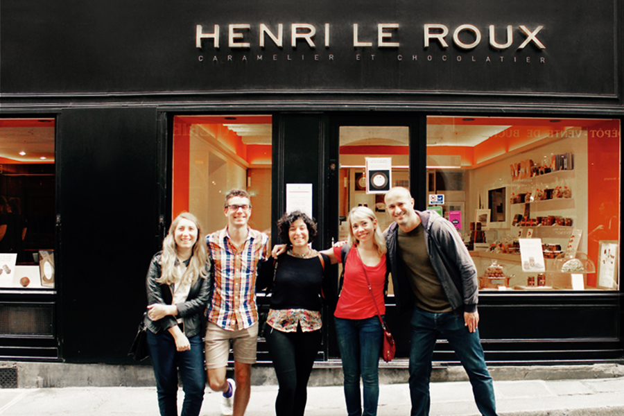Three women and two men hugging each other and posing in front of a chocolate store.