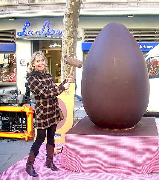 A woman who is holding a wooden hammer next to a big chocolate easter egg.