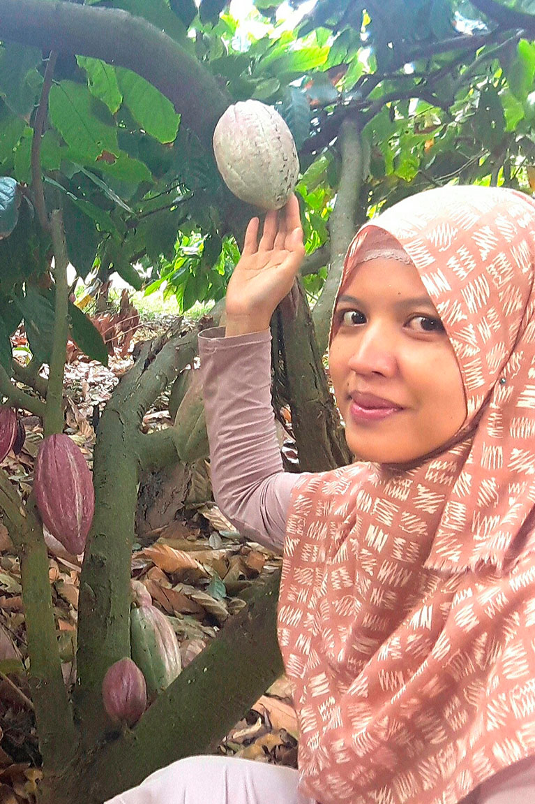 A woman posing next to a cacao tree and touching with her hand on of the cacao fruits.