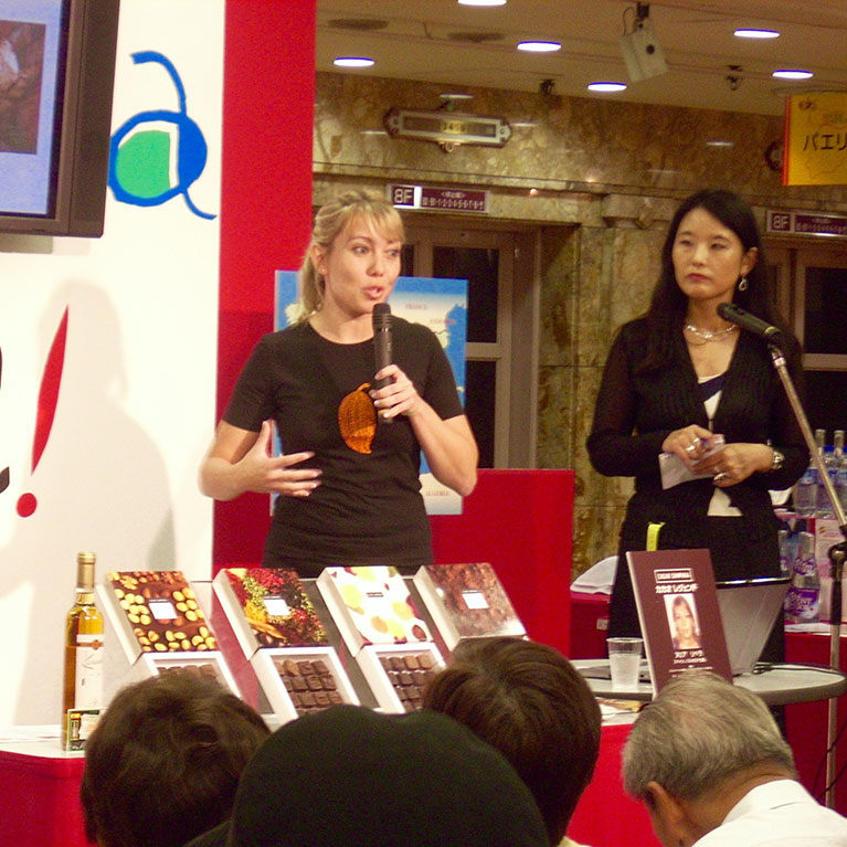 A woman talking on a microphone about chocolate origins and another woman on her left who is watching her. In front of them there are chocolaet bonbons and an alcohol bottle