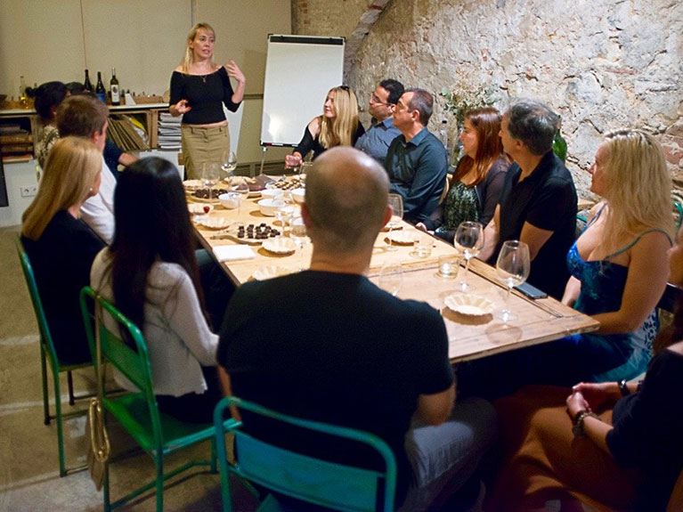 A woman giving a presentation about chocolate origins in front of a group of people who are sitting on chair around a table on which there are wine glasses and chocolate bonbons.