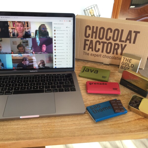 There is a laptop placed on a wooden table which is showing a Zoom meeting for chocolate tasting and on the left there are a lot of chocolate bars.