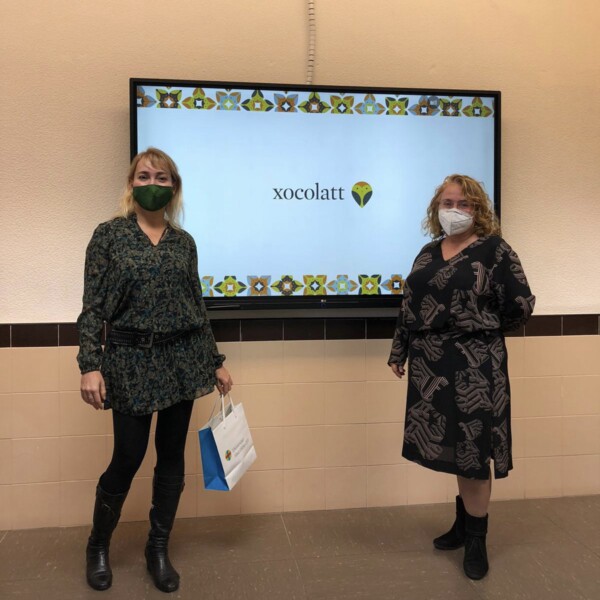 Two women with masks posing in front of an interactive board with a presentation for chocolate in a school.