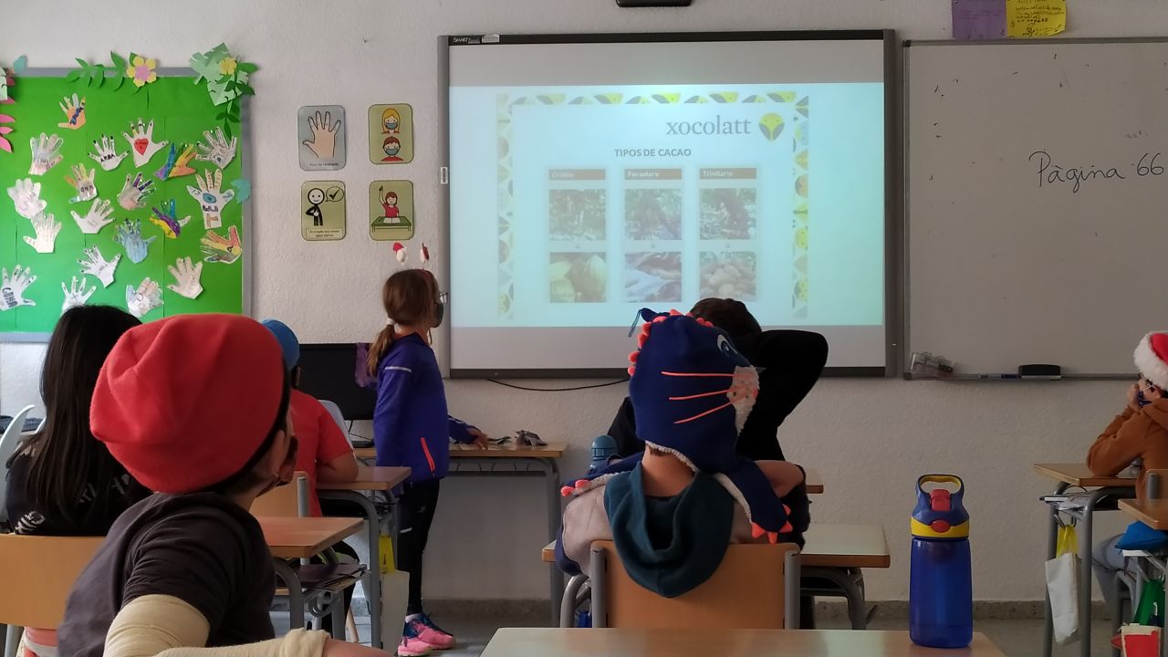 A classroom full of children sitting on their desks and there is one girl in front who is presenting a presentation about chocolate origins.